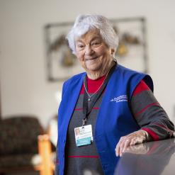 A senior woman, Theresa Acerno, wearing a blue Cheshire Medical Center volunteer vest and name badge, smiles as she leans on a counter