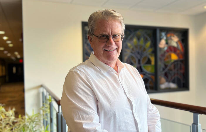 John Willard, a white man with grey hair and glasses, poses for a photo wearing a white button-down shirt.