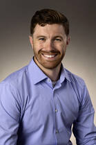 Trevor Read, DO, a white man with short brown hair and a beard, poses for a headshot wearing a blue button down shirt.