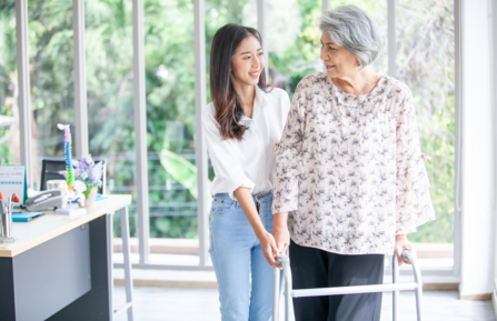 A  middle aged woman uses a walker in the kitchen as her teenaged daugter assists her with care.