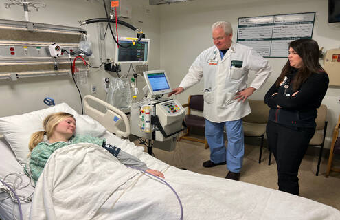 Female urse lies in a hospital bed pretending to be a patient with a male doctor and female nurse standing beside her bed