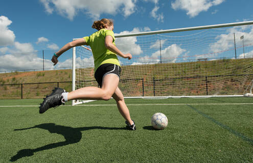 A red-headed female soccer player kicks a ball into an open net under a blue sky